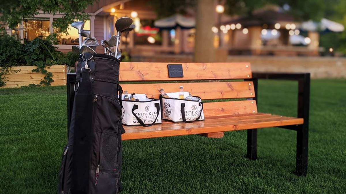 A Bar Golf League golf bag and two buckets rest on a bench by green grass near a building.