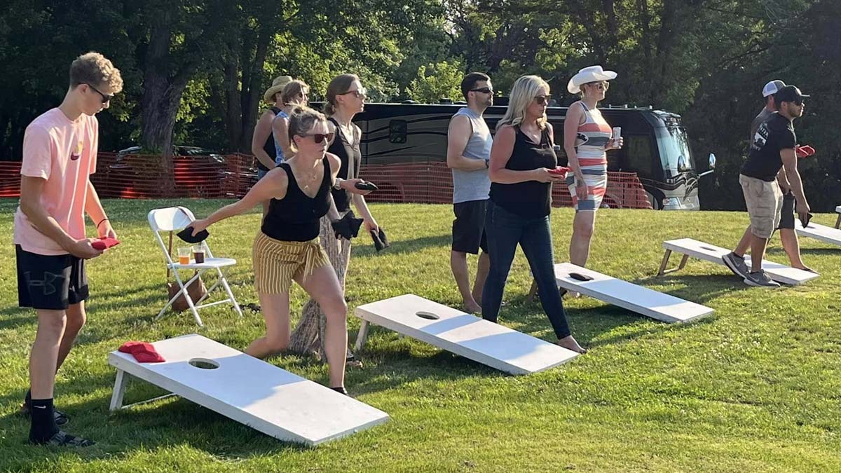 People playing cornhole at the Bean Bag Tournament Watertown, with trees and a camper nearby.