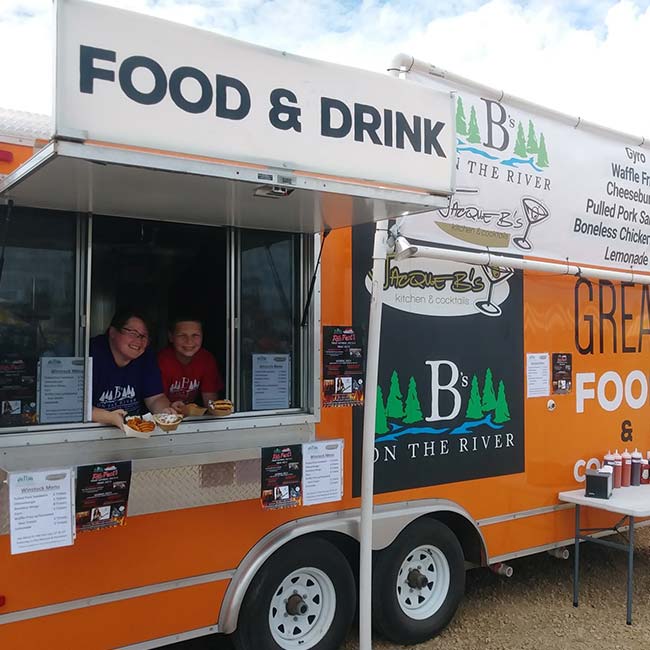 Two people serve food from a Watertown Food Trailer Catering truck labeled Bs on the River.