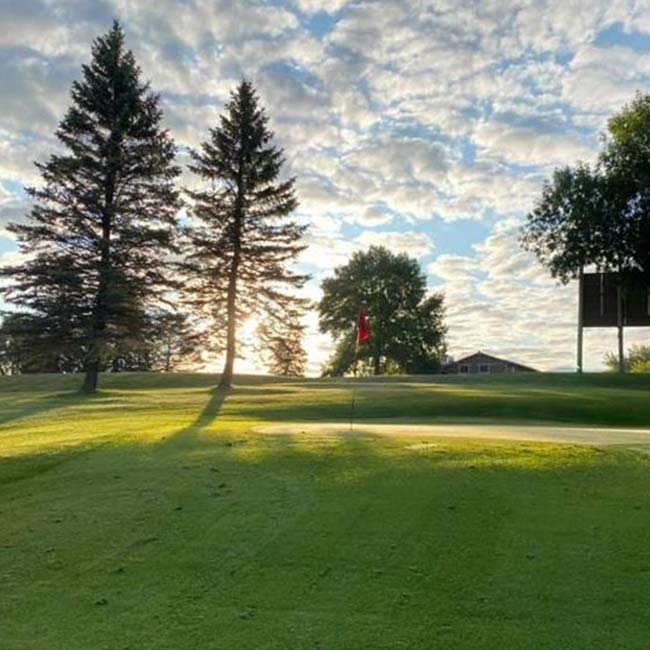 Sunrise over 9 hole golf course Watertown MN with green grass, trees, and a flagstick.