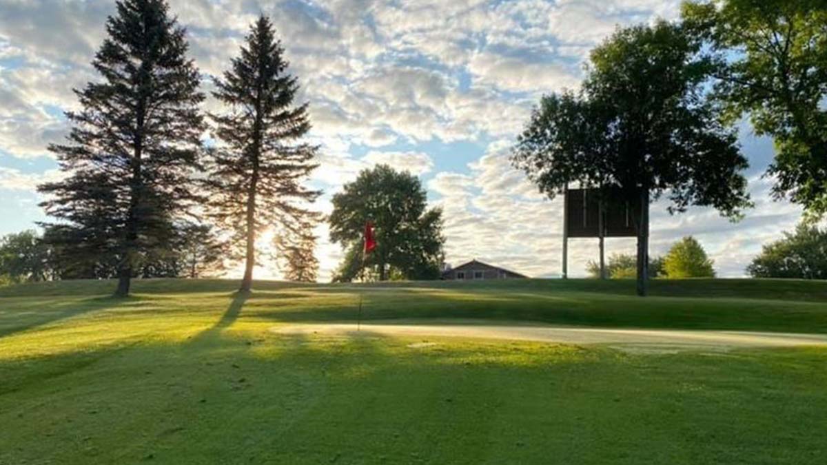 Golf course green with flagstick, trees, and Thursday Golf League at sunrise under cloudy skies.
