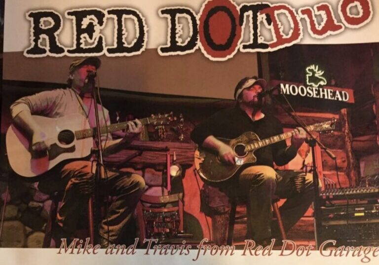 Two men playing guitars and singing on stage under a sign that says Red Dot Duo and Moosehead.